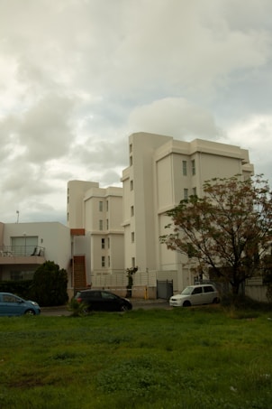 A modern white apartment building with several floors is situated behind a grassy area. In front of the building, there are three cars parked on the street. A large tree with sparse leaves stands to the right, and the sky is overcast with dense clouds.