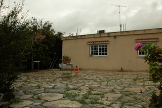 A single-story beige building with two large windows decorated with brown awnings. The foreground features a stone patio with patches of grass growing between the stones. There are various plants and trees surrounding the area, as well as a small table with a plant on it and an orange plastic stool.