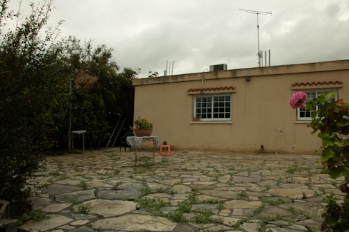 A single-story beige building with two large windows decorated with brown awnings. The foreground features a stone patio with patches of grass growing between the stones. There are various plants and trees surrounding the area, as well as a small table with a plant on it and an orange plastic stool.