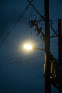 A streetlight mounted on a wooden utility pole shines brightly against a dark, cloudy sky. Several power lines extend from the pole, creating a network of cables.