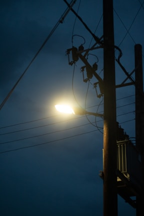 A streetlight mounted on a wooden utility pole shines brightly against a dark, cloudy sky. Several power lines extend from the pole, creating a network of cables.