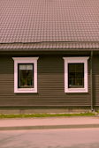 A house façade featuring a dark brown exterior with white-trimmed windows. The roof is covered in patterned shingles, and two windows are visible along the wall, each with visible interior plants. Below, there is a small strip of grass separating the building from the pavement.