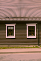 A house façade featuring a dark brown exterior with white-trimmed windows. The roof is covered in patterned shingles, and two windows are visible along the wall, each with visible interior plants. Below, there is a small strip of grass separating the building from the pavement.