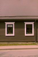 A house façade featuring a dark brown exterior with white-trimmed windows. The roof is covered in patterned shingles, and two windows are visible along the wall, each with visible interior plants. Below, there is a small strip of grass separating the building from the pavement.