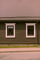 A house façade featuring a dark brown exterior with white-trimmed windows. The roof is covered in patterned shingles, and two windows are visible along the wall, each with visible interior plants. Below, there is a small strip of grass separating the building from the pavement.