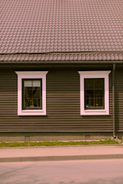A house façade featuring a dark brown exterior with white-trimmed windows. The roof is covered in patterned shingles, and two windows are visible along the wall, each with visible interior plants. Below, there is a small strip of grass separating the building from the pavement.