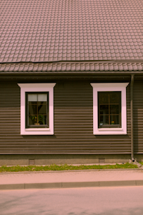 A house façade featuring a dark brown exterior with white-trimmed windows. The roof is covered in patterned shingles, and two windows are visible along the wall, each with visible interior plants. Below, there is a small strip of grass separating the building from the pavement.