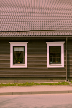 A house façade featuring a dark brown exterior with white-trimmed windows. The roof is covered in patterned shingles, and two windows are visible along the wall, each with visible interior plants. Below, there is a small strip of grass separating the building from the pavement.