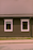 A house façade featuring a dark brown exterior with white-trimmed windows. The roof is covered in patterned shingles, and two windows are visible along the wall, each with visible interior plants. Below, there is a small strip of grass separating the building from the pavement.