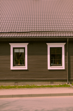 A house façade featuring a dark brown exterior with white-trimmed windows. The roof is covered in patterned shingles, and two windows are visible along the wall, each with visible interior plants. Below, there is a small strip of grass separating the building from the pavement.