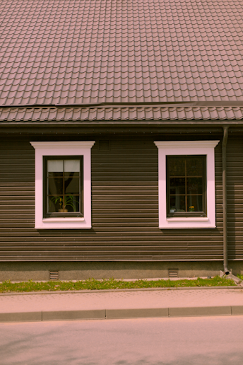 A house façade featuring a dark brown exterior with white-trimmed windows. The roof is covered in patterned shingles, and two windows are visible along the wall, each with visible interior plants. Below, there is a small strip of grass separating the building from the pavement.