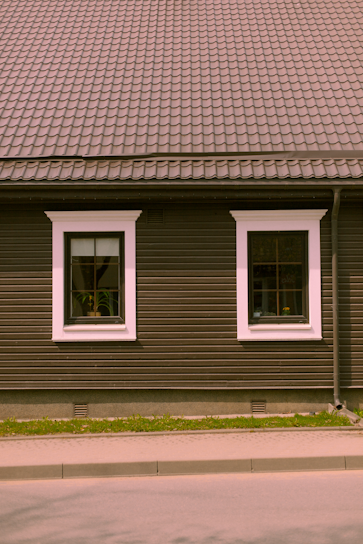 A house façade featuring a dark brown exterior with white-trimmed windows. The roof is covered in patterned shingles, and two windows are visible along the wall, each with visible interior plants. Below, there is a small strip of grass separating the building from the pavement.