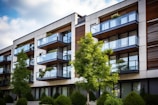 Modern apartment building with large glass balconies and a clean, linear design. The facade combines elements of white concrete and dark wood, harmoniously blending with the greenery of neatly trimmed bushes and trees in the foreground.