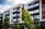 Modern apartment building with large glass balconies and a clean, linear design. The facade combines elements of white concrete and dark wood, harmoniously blending with the greenery of neatly trimmed bushes and trees in the foreground.