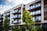 Modern apartment building with large glass balconies and a clean, linear design. The facade combines elements of white concrete and dark wood, harmoniously blending with the greenery of neatly trimmed bushes and trees in the foreground.
