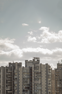 A row of tall, rectangular buildings with numerous balconies and windows under a cloudy sky. The architecture appears worn, with a vintage aesthetic. A sign that seems to read 'VILNIUS' is mounted on top of one of the buildings.