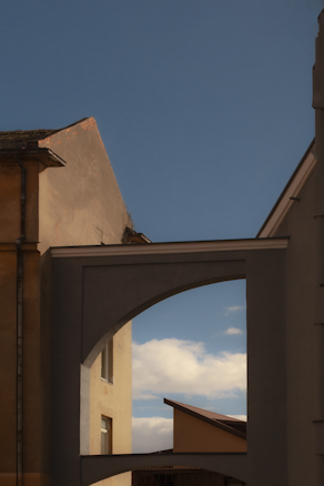 An architectural scene with buildings forming geometric shapes against a blue sky. An archway frames a view of other structures and some clouds are visible. The walls display earthy tones with hints of shadows and light.