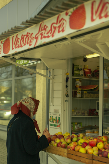 A person wearing a headscarf is standing at an outdoor market stall filled with apples. The stall has various canned goods displayed, and there is signage with fruit imagery. The scene appears to be in an urban setting with a building in the background.