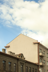 A section of urban architecture is presented, featuring a beige, flat-roofed building with minimalistic design elements. Adjacent is an older, ornate structure with visible details like chimneys and decorative molding. The sky is partly cloudy with patches of blue, adding contrast to the scene.