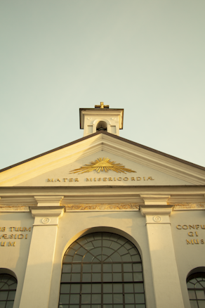 A close-up of a classical church facade featuring a prominent triangular pediment with the inscription 'Mater Misericordiae'. The architectural style includes columns and an arched window below. The sunlight casts a warm glow, enhancing the golden details on the facade.