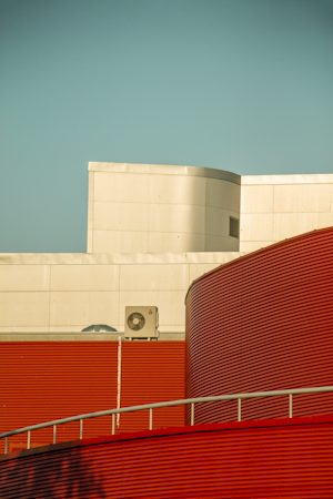 The image features a section of a modern architectural building with a contrast of smooth and corrugated surfaces. The structure includes a light beige, smooth, curving area at the top, juxtaposed against a bold red, corrugated facade in the lower half, with horizontal lines. An industrial air conditioning unit is visible towards the center of the white section.