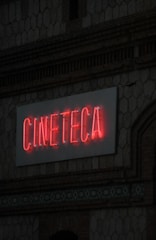 A neon sign with the word 'CINETECA' is illuminated in red against a textured wall. The architectural details include a patterned design and a brickwork facade.