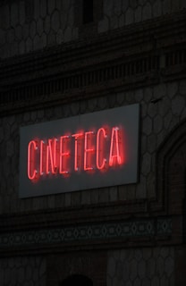 A neon sign with the word 'CINETECA' is illuminated in red against a textured wall. The architectural details include a patterned design and a brickwork facade.