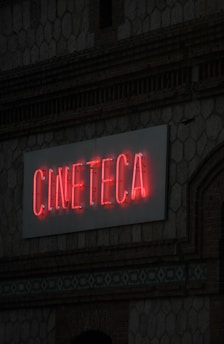 A neon sign with the word 'CINETECA' is illuminated in red against a textured wall. The architectural details include a patterned design and a brickwork facade.