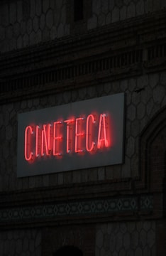A neon sign with the word 'CINETECA' is illuminated in red against a textured wall. The architectural details include a patterned design and a brickwork facade.