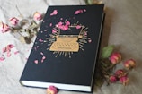 A hardcover notebook lies on a cream-colored textile surface, adorned with a golden typewriter design on the cover. Scattered around and on the notebook are dried pink rose petals and a few dried roses, adding a touch of elegance and vintage charm.