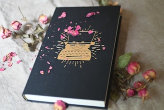 A hardcover notebook lies on a cream-colored textile surface, adorned with a golden typewriter design on the cover. Scattered around and on the notebook are dried pink rose petals and a few dried roses, adding a touch of elegance and vintage charm.