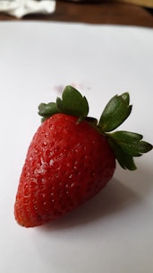 A fresh, ripe strawberry with a vibrant red color is positioned on a white surface. The strawberry's seeds are clearly visible, and its green leaves appear crisp and healthy.