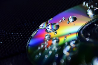 Close-up of a CD with water droplets scattered on its surface. The light creates a spectrum of rainbow colors on the CD, enhancing its reflective properties. The background is a textured dark surface which contrasts with the vibrant colors.