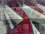 Geometric glass panels create a complex pattern with reflections of an urban landscape. Buildings appear distorted in the glass, providing an abstract and modern architectural view.