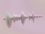 A 3D abstract form resembling a sound wave pattern is displayed against a soft pink and beige gradient background. The wave-like structure consists of smooth, rippling layers that give it a sense of depth and fluidity.