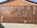 A large wall display featuring an array of wooden plaques with Chinese characters engraved on them. The plaques vary in size and are arranged in a seemingly random pattern against a backdrop of tiles. The bright sunlight casts shadows, highlighting the texture and depth of the engravings.