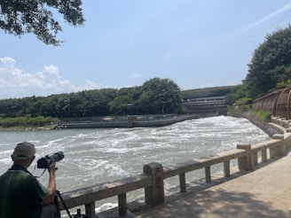 A person with a telescope is observing a wide, flowing river surrounded by lush greenery. A concrete railing separates the observer from the water. In the background, a bridge and dense forest create a lush setting. The sky is clear with a few clouds, suggesting a sunny day.