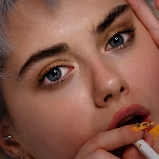A close-up of a person's face highlighting their expressive eyes and detailed makeup. The eye shadow is a subtle gold, and the eyebrows are bold and defined. The person has pale skin and short, light hair. Their nails are painted with intricate designs in gold and black, and they are holding what appears to be a cigarette near their lips.