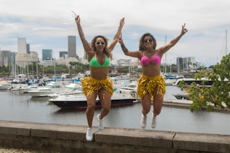 Two women wearing colorful bikini tops and gold fringed skirts are jumping in the air, showing excitement and energy. Behind them, a marina is filled with various boats and yachts, setting an urban background with a city skyline visible in the distance.