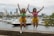 Two women wearing colorful bikini tops and gold fringed skirts are jumping in the air, showing excitement and energy. Behind them, a marina is filled with various boats and yachts, setting an urban background with a city skyline visible in the distance.