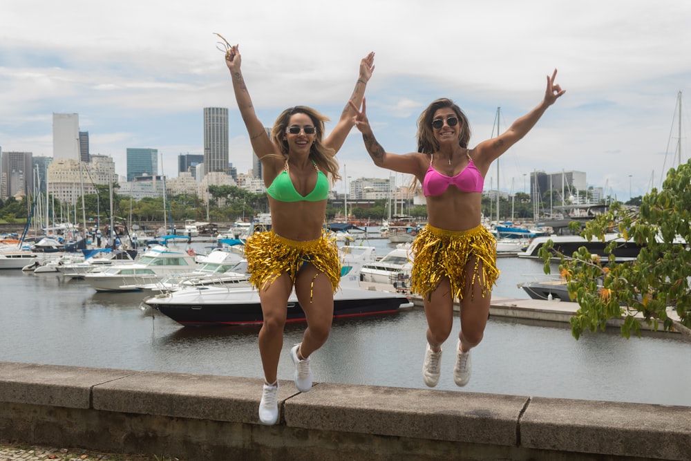two women in bikinis jumping in front of a marina