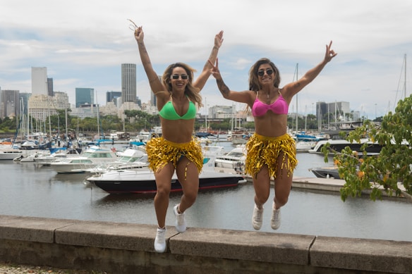 Two women wearing colorful bikini tops and gold fringed skirts are jumping in the air, showing excitement and energy. Behind them, a marina is filled with various boats and yachts, setting an urban background with a city skyline visible in the distance.
