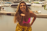 A woman with long hair, wearing a bright pink bikini top and a shiny yellow skirt, stands near a marina with various boats in the background. She is smiling and posing with one hand on her hip and the other hand raised towards her mouth.