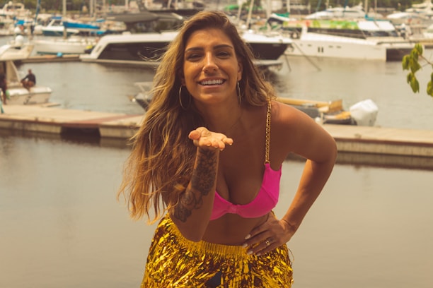 A woman with long hair, wearing a bright pink bikini top and a shiny yellow skirt, stands near a marina with various boats in the background. She is smiling and posing with one hand on her hip and the other hand raised towards her mouth.