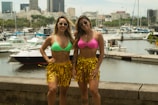 Two women wearing colorful bikini tops and gold fringed skirts are posing by a waterfront. They are both wearing sunglasses. Several boats and yachts are docked in the marina behind them, and a city skyline is visible in the background.