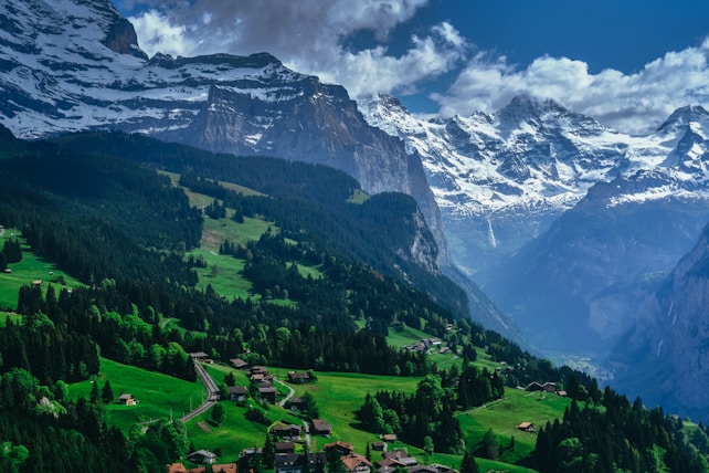 A picturesque landscape features a lush green valley with scattered houses, surrounded by dense forests. Towering snow-capped mountains rise dramatically in the background, under a partly cloudy sky, creating a striking contrast between the verdant foreground and the rugged peaks.