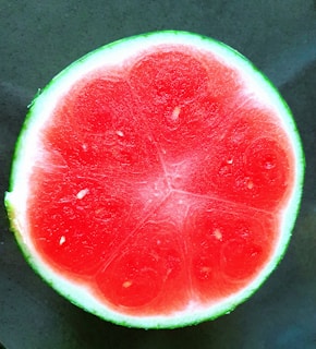 A cross-section of a vibrant red watermelon with a thin green rind bordering the bright, juicy interior. The texture is smooth with a few seeds visible within the pulp.