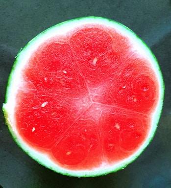 A cross-section of a vibrant red watermelon with a thin green rind bordering the bright, juicy interior. The texture is smooth with a few seeds visible within the pulp.