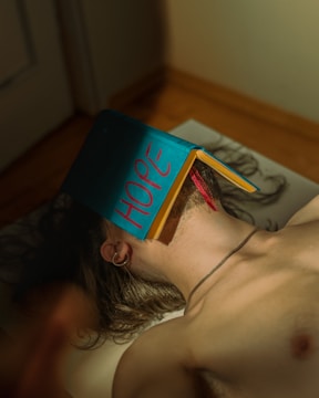 A person is lying on their back with a blue book titled 'HOPE' covering their face. The person has long hair, and the scene is set indoors, illuminated by soft lighting. The book's spine is facing upwards, and a pink ribbon is visible hanging from the book. The person is wearing an ear piercing and a thin necklace.