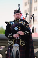 A man dressed in traditional Scottish attire plays a set of bagpipes. He wears a kilt with a tartan pattern and a matching sash, along with a dark jacket adorned with silver buttons. The background shows a blurred outdoor setting with a building and sky visible.